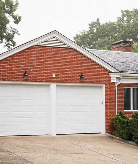 Exterior view of a brick house with two white garage doors. The roof is shingled, and there are green bushes along the side, conveying a neat and tidy appearance.