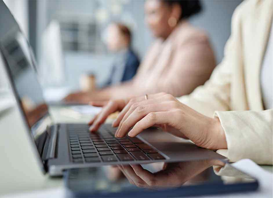 Close-up of hands typing on a laptop in a modern office. Two blurred colleagues work in the background. The scene conveys focus and productivity.