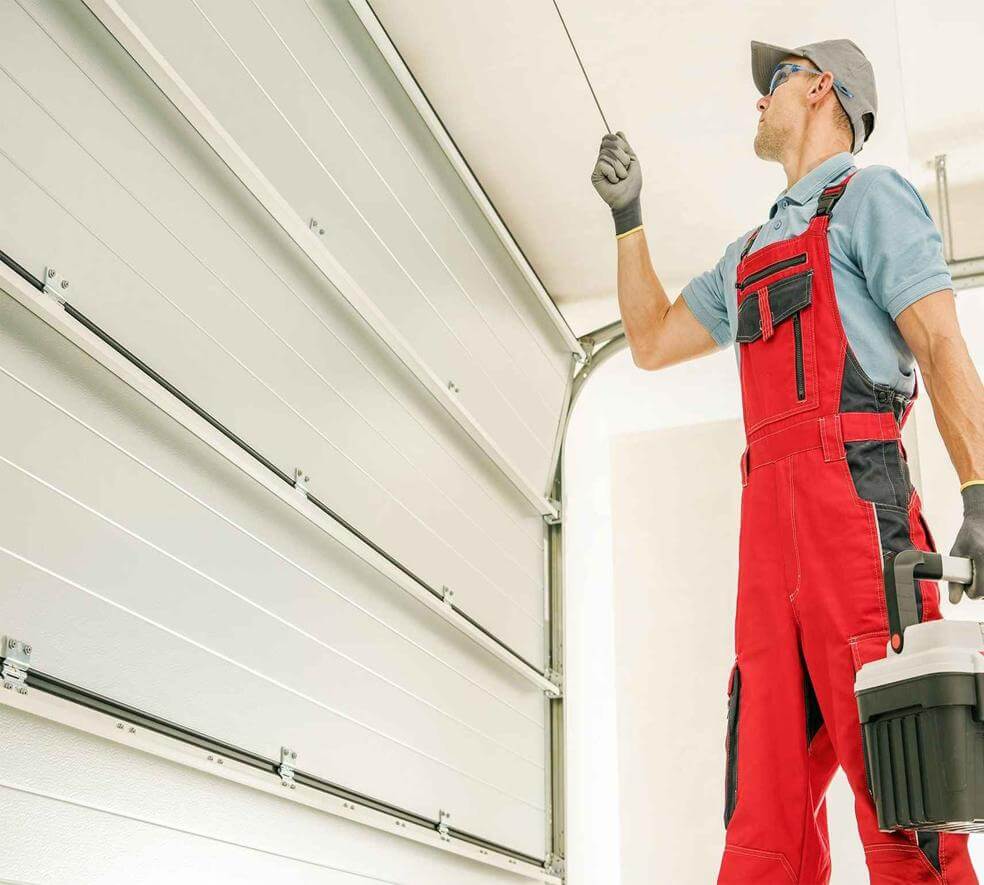 A man in red overalls inspects a garage door mechanism. He holds a toolbox, wearing gloves and a cap. The scene conveys focus and professionalism.