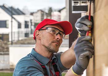 A focused construction worker wearing a red cap and safety goggles uses a screwdriver to adjust an outdoor panel. Modern buildings are in the background.