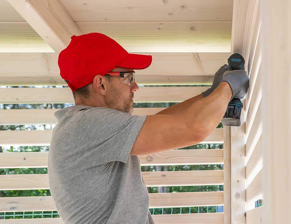 A person in a red cap uses a cordless drill on light wooden planks, wearing safety glasses and gloves, conveying focus and craftsmanship in a workshop setting.