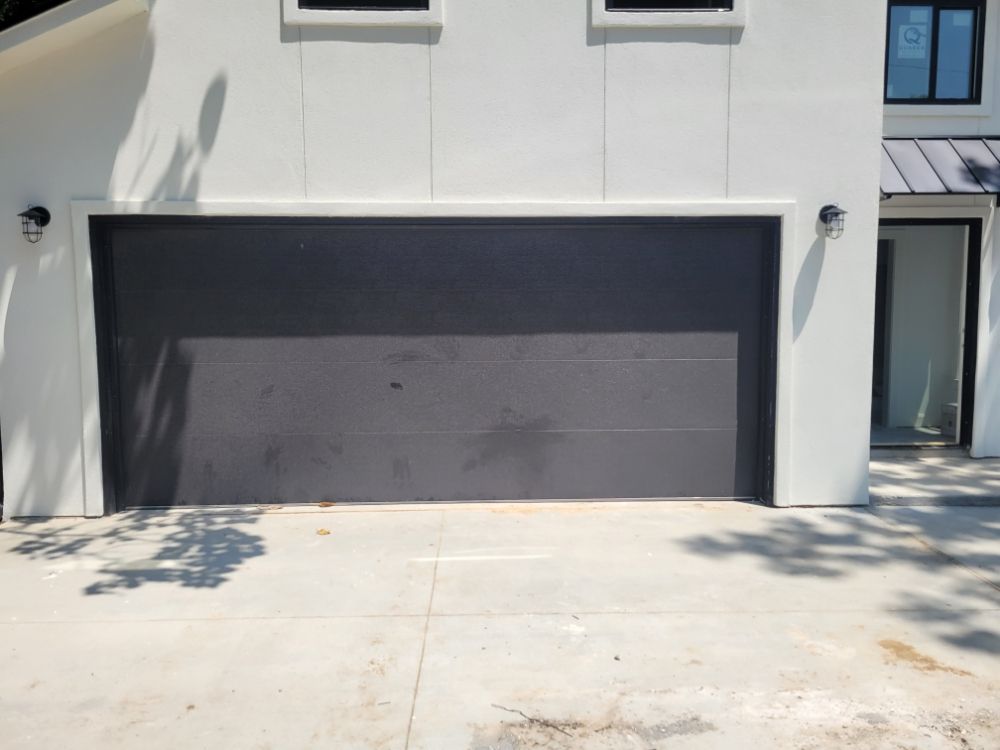 Modern house exterior with a smooth black garage door. The building is white with minimalistic style, and shadows from nearby trees play on the driveway.