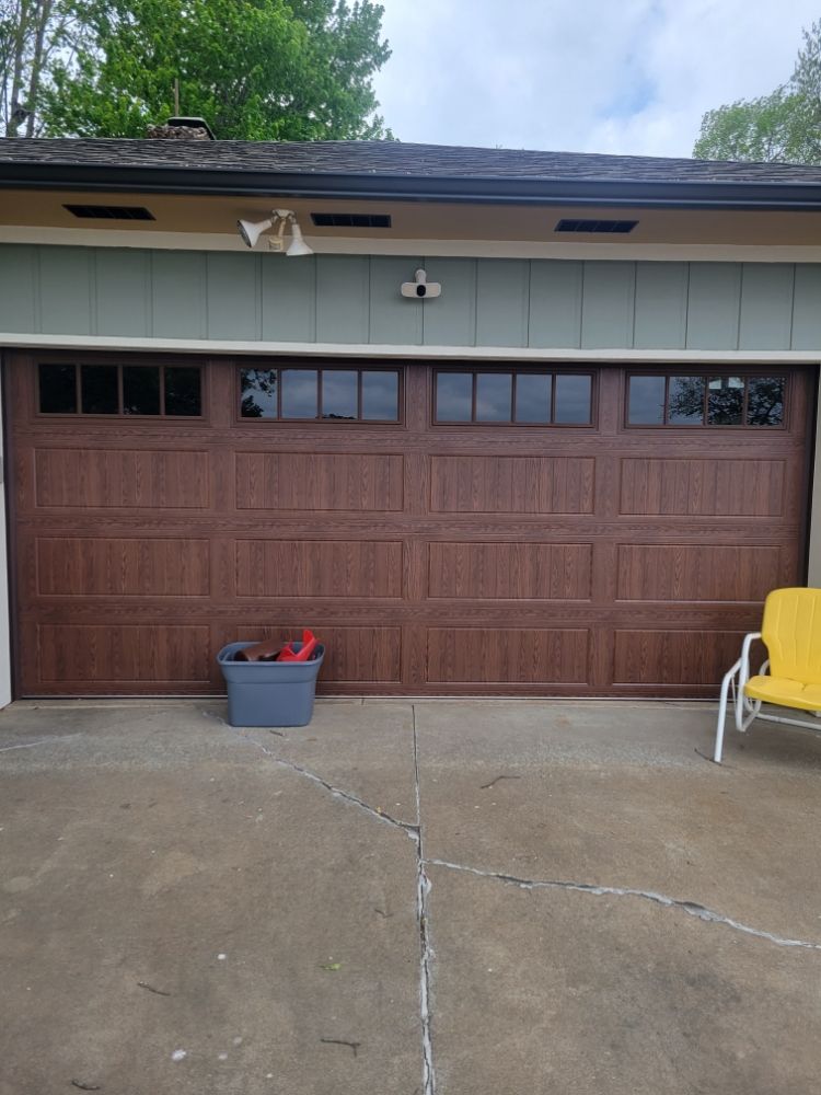 Closed brown garage door with windows at the top, a gray tote with items on the concrete driveway, and a yellow chair on the right. Greenery in the background.