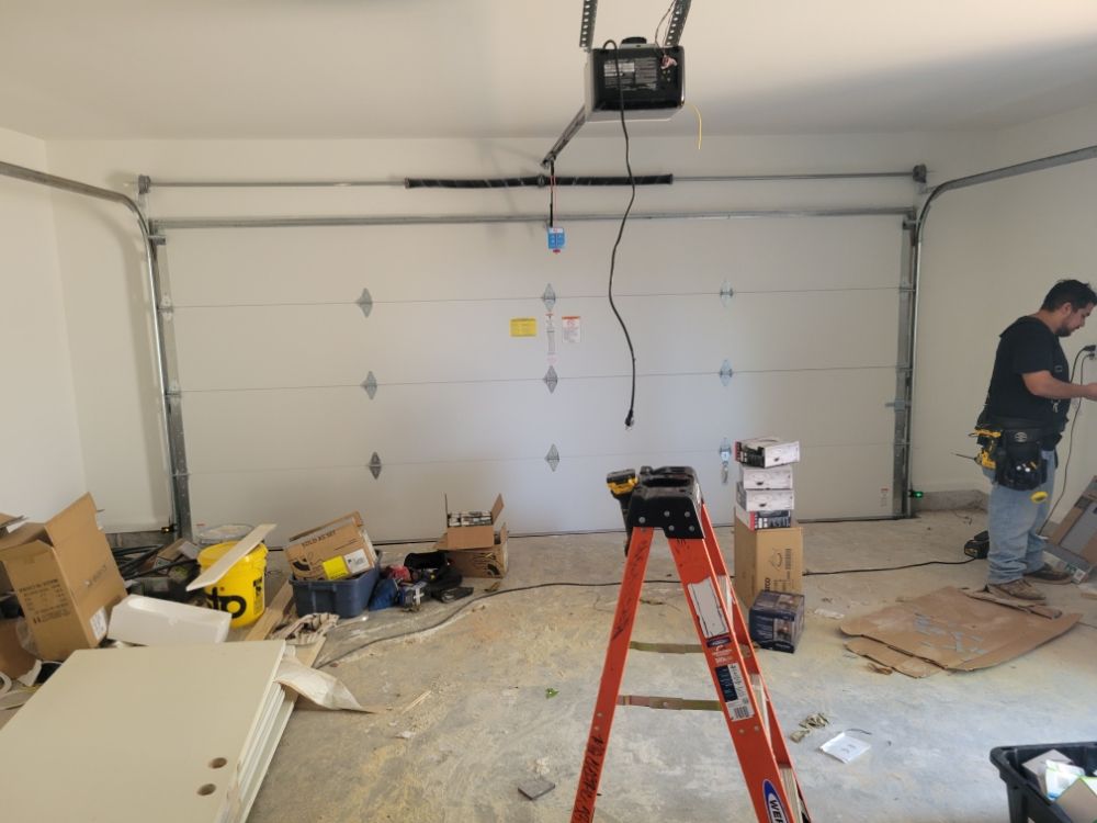 A cluttered garage with a white door and overhead opener. A red ladder stands in the center amidst scattered tools, boxes, and a working person on the right.