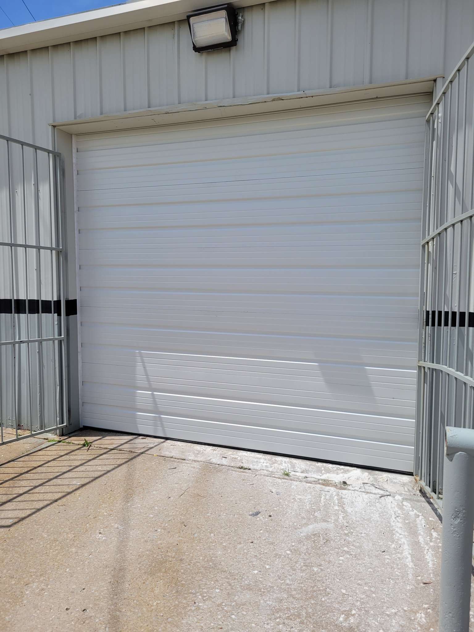A closed white garage door on a metal building is illuminated by sunlight. The concrete ground is slightly weathered, with faint shadows visible.